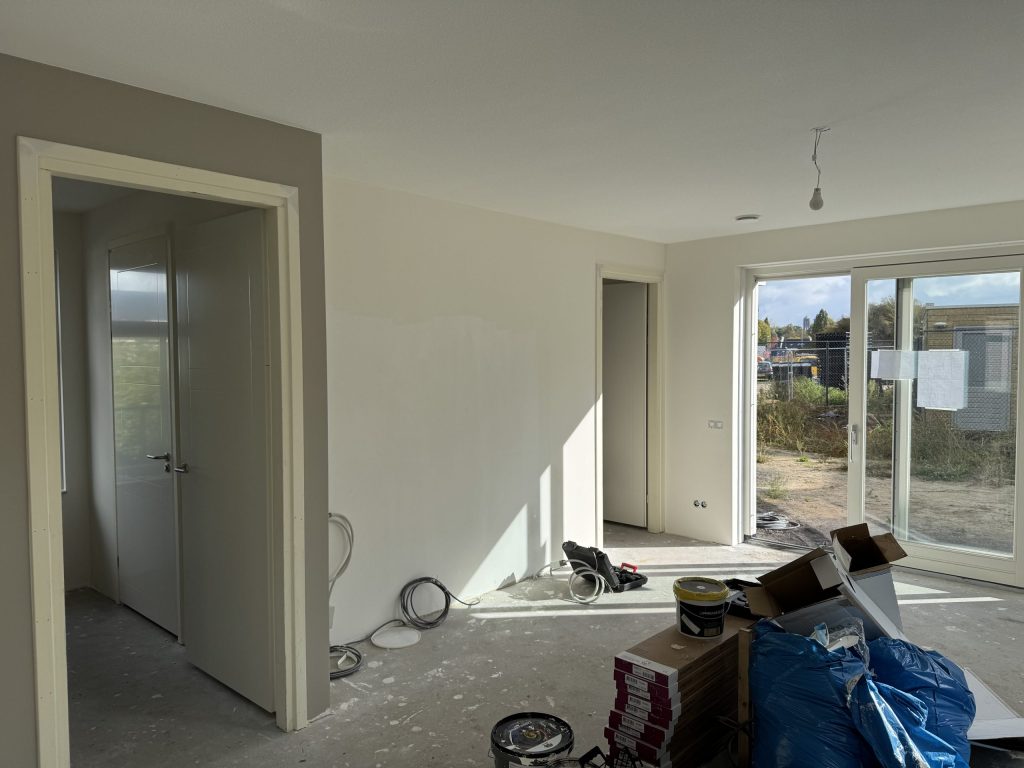 Woonkamer van een net opgeleverde nieuwbouwwoning met verfemmers, dozen en kabels op de betonvloer